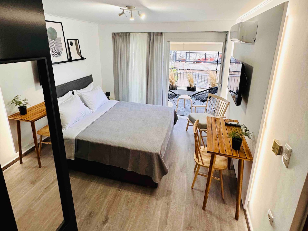 The mini-suite features a queen-size bed with clean linens, complemented by a spacious wardrobe. A cozy sitting area with wooden furniture is highlighted by large windows that lead to a private balcony. A wall-mounted smart TV adds to the modern appeal.