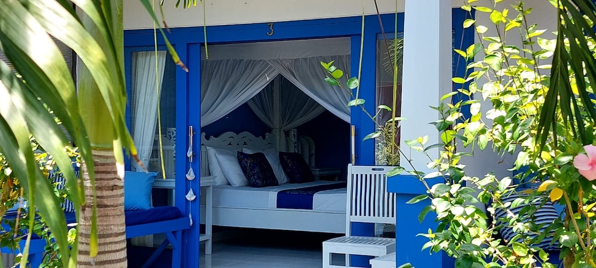 A private terrace is framed by lush greenery and vibrant flowers, leading to a stylish bedroom featuring a spacious bed draped in white curtains. Blue accents on the exterior and furniture create a fresh atmosphere, enhancing the connection to the surrounding nature.