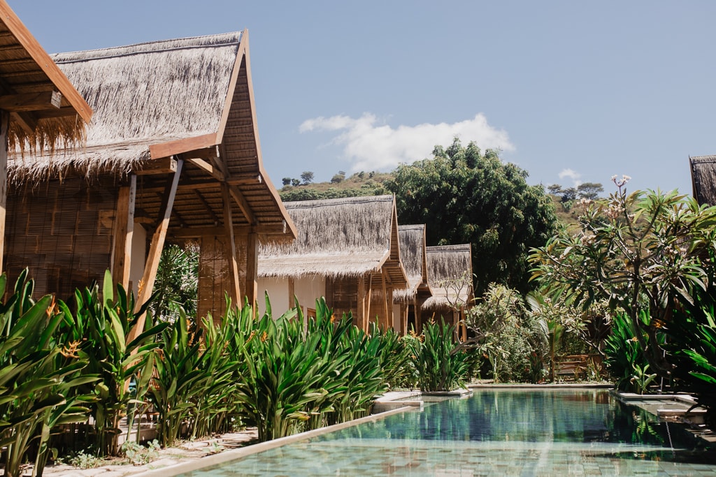 A tranquil scene is presented with a line of bungalows featuring thatched roofs, set against a backdrop of lush greenery. A serene swimming pool reflects the blue sky above, flanked by vibrant tropical plants, enhancing the natural beauty of the environment.