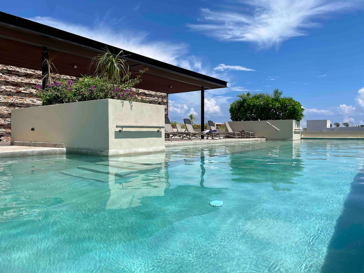 A pool area is highlighted, featuring a crystal-clear water surface reflecting the blue sky. Lush greenery decorates planters, and multiple lounge chairs are arranged along the poolside, creating a serene atmosphere. The modern structure provides a shaded terrace above.