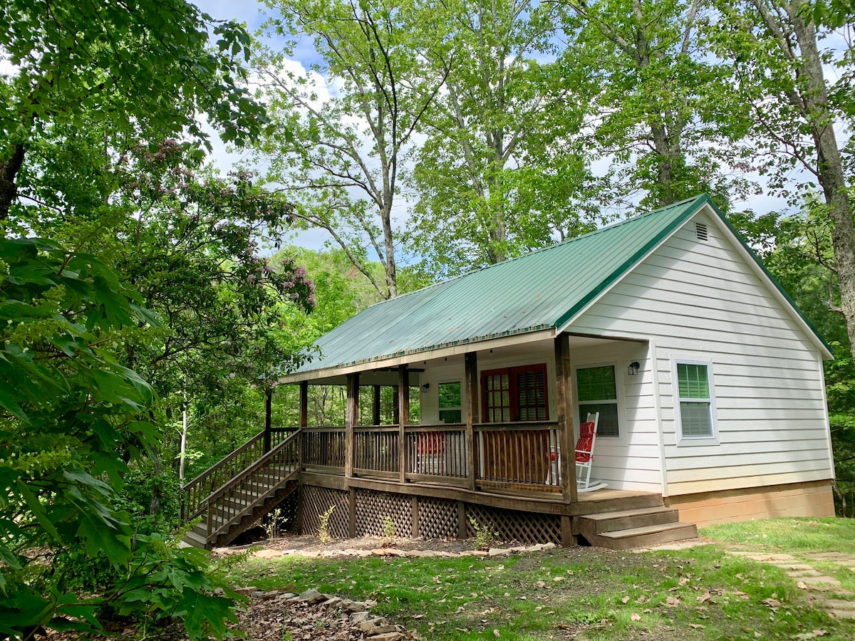 A cabin with a green metal roof is nestled among lush trees. The covered porch features wooden railings and multiple red rocking chairs, inviting relaxation. Natural surroundings enhance the tranquility of the location, with a pathway leading to the entrance, accessible for guests.