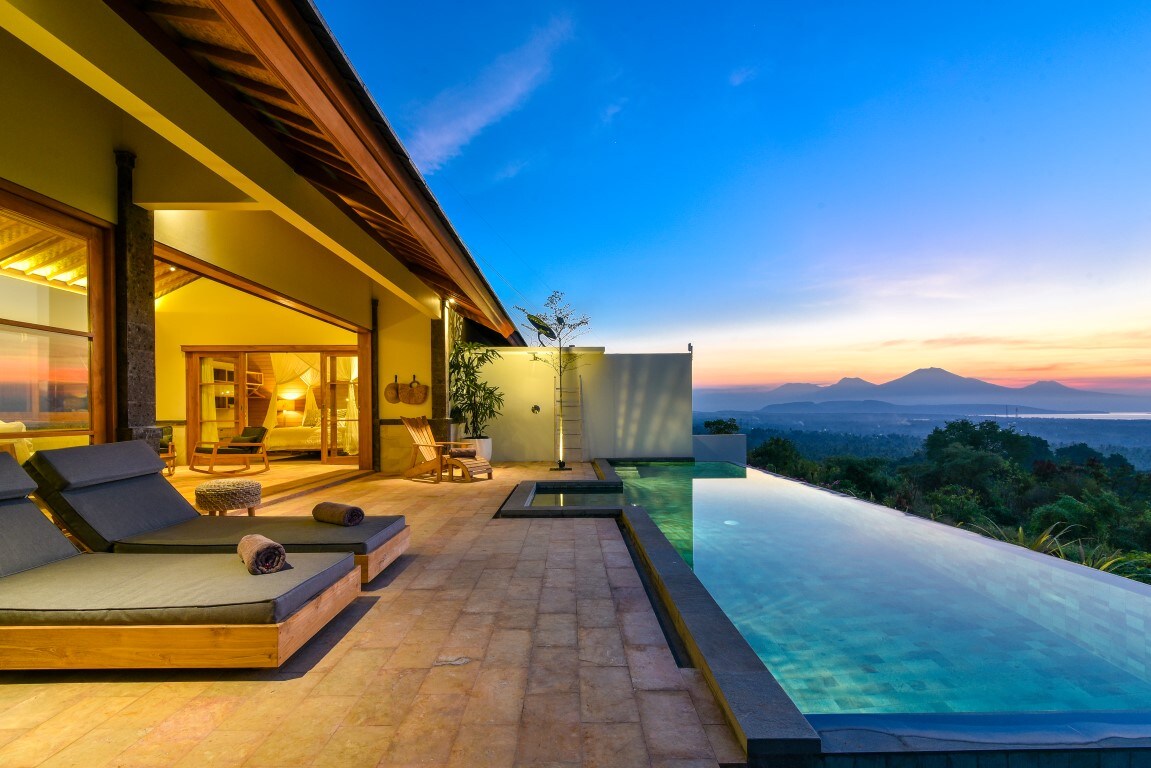 An inviting outdoor area features a spacious infinity pool reflecting the evening sky, surrounded by comfortable lounge chairs. The open living space showcases large sliding glass doors leading to stunning views of distant mountains and a colorful sunset.