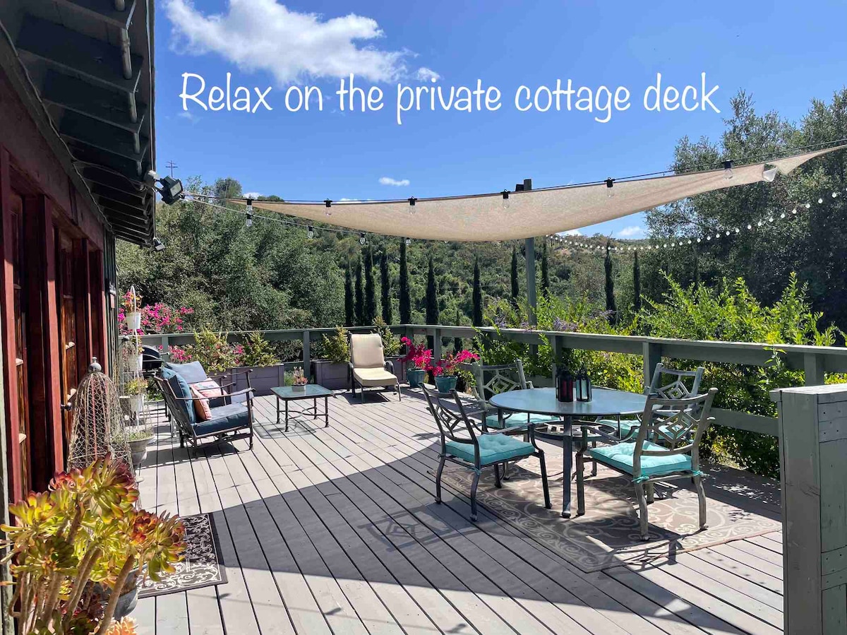 The private cottage deck features comfortable seating arrangements, including a dining table surrounded by chairs and lounge chairs positioned for relaxation. A shade structure provides protection from the sun, while greenery surrounds the space, creating a serene outdoor environment.