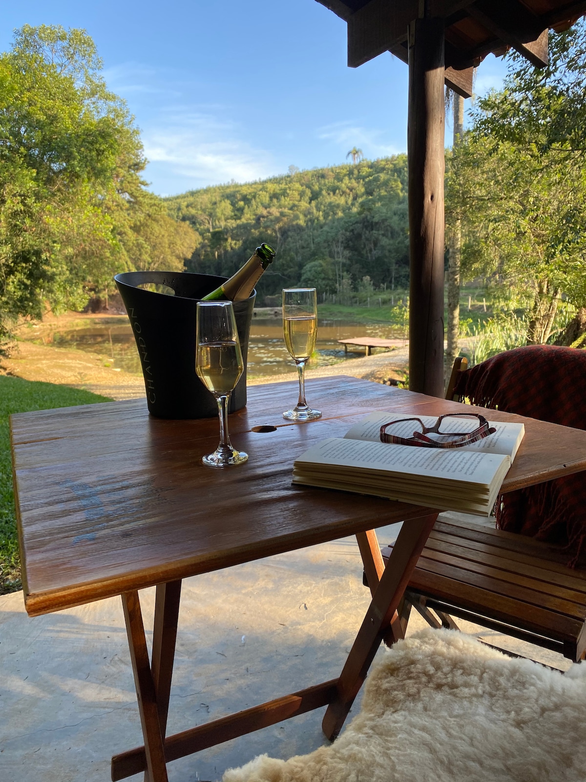 A wooden table is set with two glasses filled with sparkling beverage next to an ice bucket and a book, accompanied by a pair of reading glasses. The table is positioned on a covered patio, offering a view of lush greenery and a serene water feature in the background.