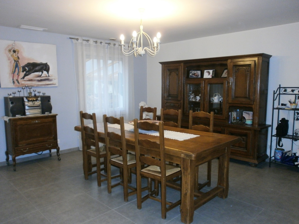 A large dining area features a rustic wooden table surrounded by eight chairs. A chandelier hangs above the table, and a sideboard and shelving unit display decorative items. Natural light filters through the window, which is dressed with sheer curtains.