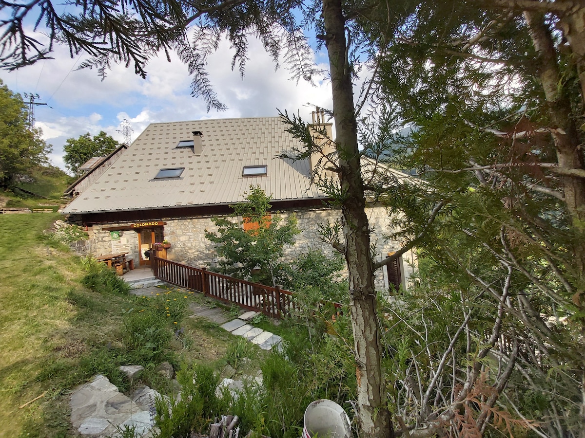 A natural landscape surrounds the rustic stone building, partially obscured by trees. The structure features a sloped roof and a welcoming entrance with a wooden railing. A pathway lined with greenery leads toward the entrance, inviting visitors to explore the tranquil setting.