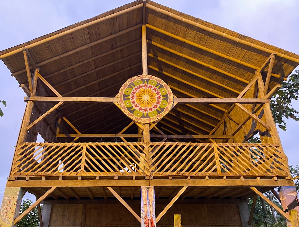 A two-story structure constructed from bamboo and wood is showcased, featuring a large circular mural with vibrant colors at the center. The open balcony with wooden railings allows for airflow and provides views of the surrounding greenery.