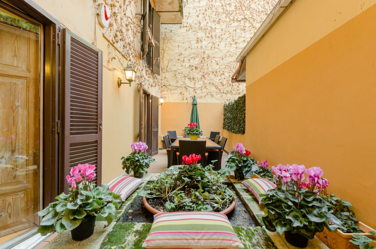 A charming patio is enriched with blooming flowers in vibrant shades of pink, surrounded by greenery. A dining table with chairs is placed under a shaded umbrella, inviting al fresco meals. Walls adorned with climbing plants provide a tranquil, secluded atmosphere.