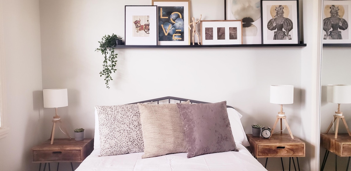 A cozy bedroom showcases a spacious bed adorned with decorative pillows. On either side, wooden nightstands hold table lamps. Above the bed, a shelf displays framed artwork and a plant, adding a touch of greenery to the neutral wall.