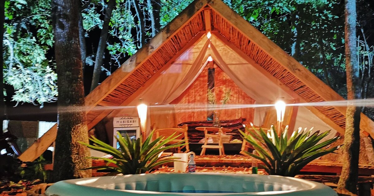 A charming tent pavilion is surrounded by lush greenery, featuring soft lighting that highlights its natural wooden structure. In front, a cozy seating area is visible, while a circular pool adds a refreshing touch to the serene outdoor environment.