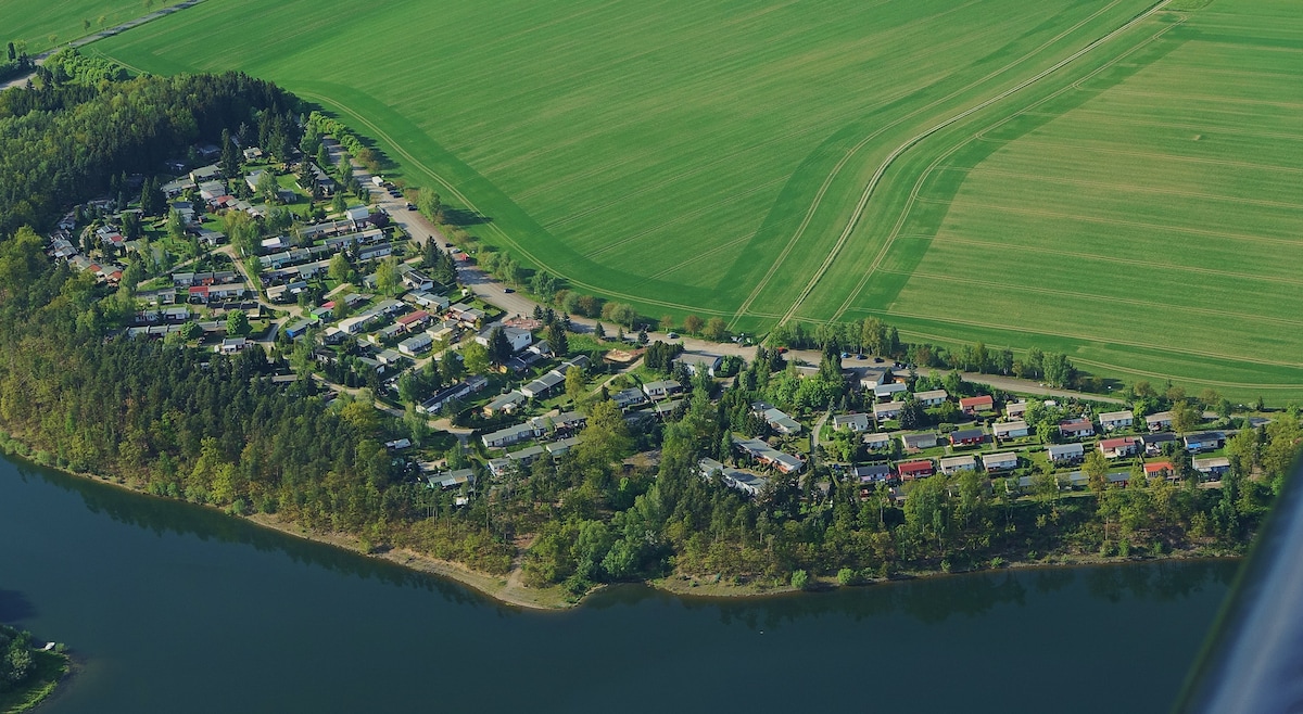 An aerial view captures a vacation complex nestled along the shore of a lake, surrounded by lush greenery and open fields. Windrows and roads weave through the development, which features various residential buildings with distinct roofs and colorful facades.