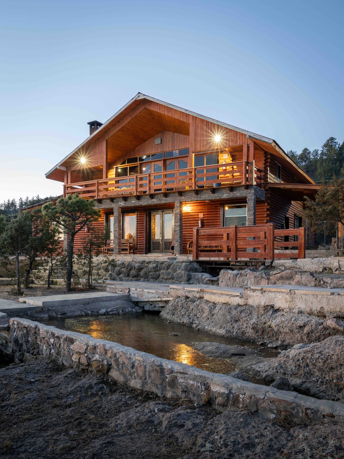 A rustic wooden cabin is set amidst a natural landscape at dusk. The multi-level structure features a balcony with wooden railings, and warm light glows from the windows. A stone pathway leads to a water feature, enhancing the outdoor setting.