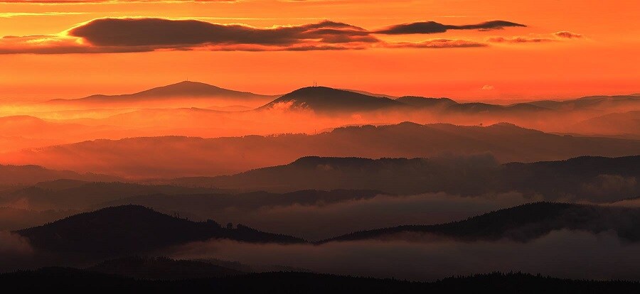 A panoramic view of rolling hills is presented under a vibrant sunset sky. Layers of purple and orange hues merge with soft clouds, creating a serene landscape that extends into the distance, showcasing the natural beauty of the surrounding mountains.
