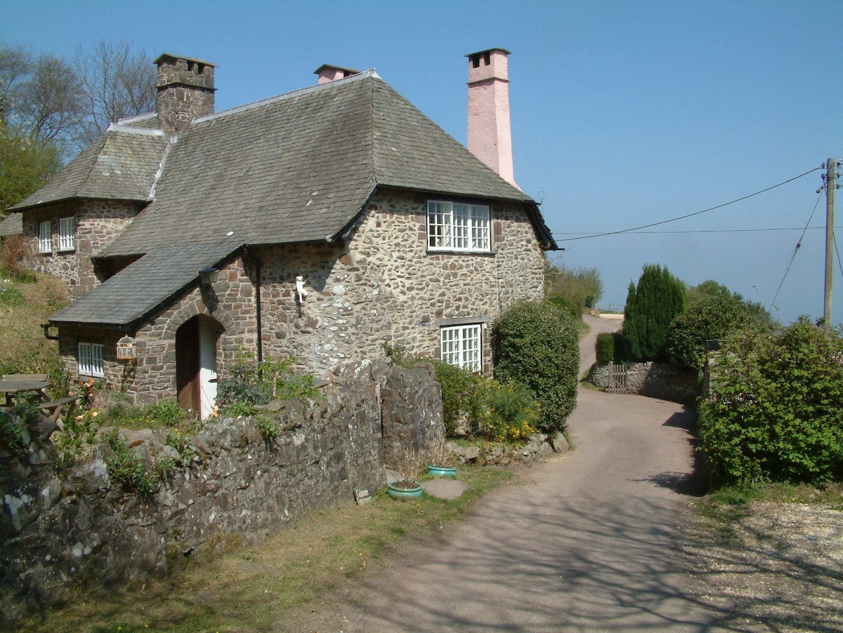 A traditional stone cottage is nestled amid greenery, featuring a charming slate roof and multiple chimneys. The entrance is framed by flowering plants and a winding path leads from the cottage to a gravel driveway, enhancing the rural setting.
