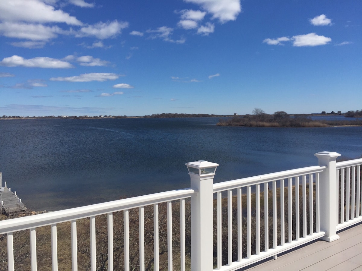A waterfront view features calm blue waters extending towards the horizon, framed by a white railing. The sky above is dotted with fluffy clouds, complementing the serene landscape of the pond and surrounding vegetation.