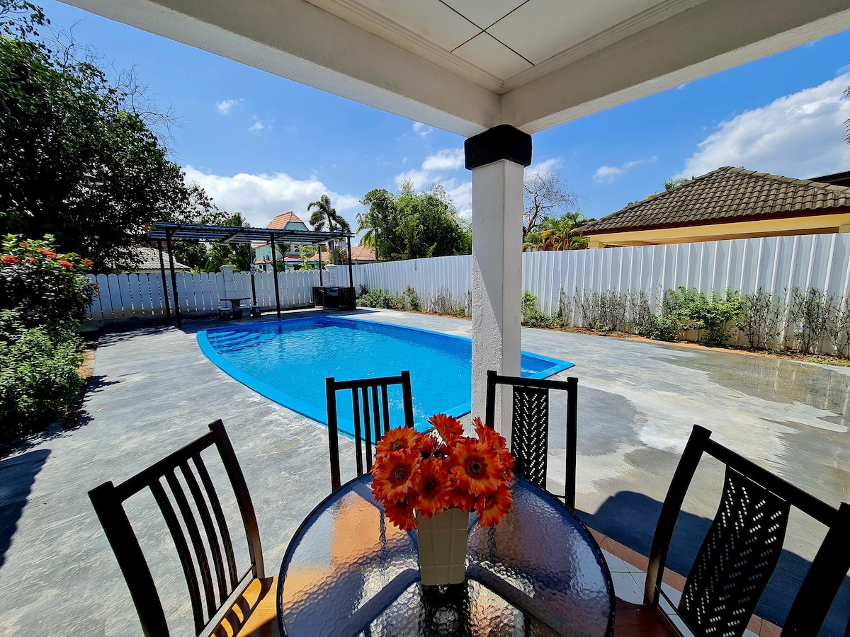 An outdoor seating area includes a round glass table surrounded by six chairs. A bouquet of bright flowers is centered on the table. Behind, a swimming pool reflects blue skies, while a white fence and lush greenery frame the scene.