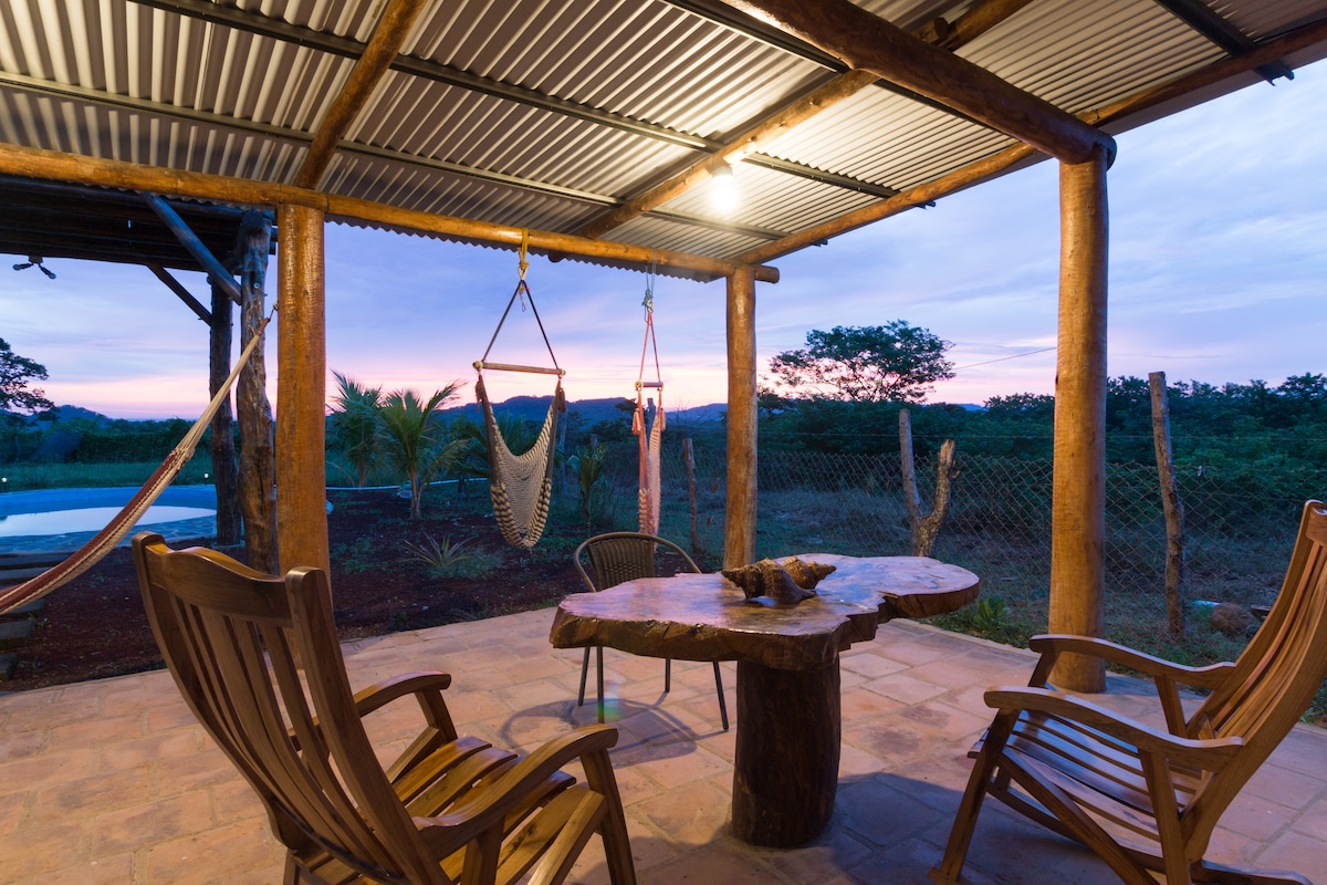 A covered patio area features a rustic wooden table surrounded by two chairs. Two hammock chairs are suspended nearby. The tranquil outdoor setting includes a view of the landscaped yard and swimming pool, with a soft sunset illuminating the sky.