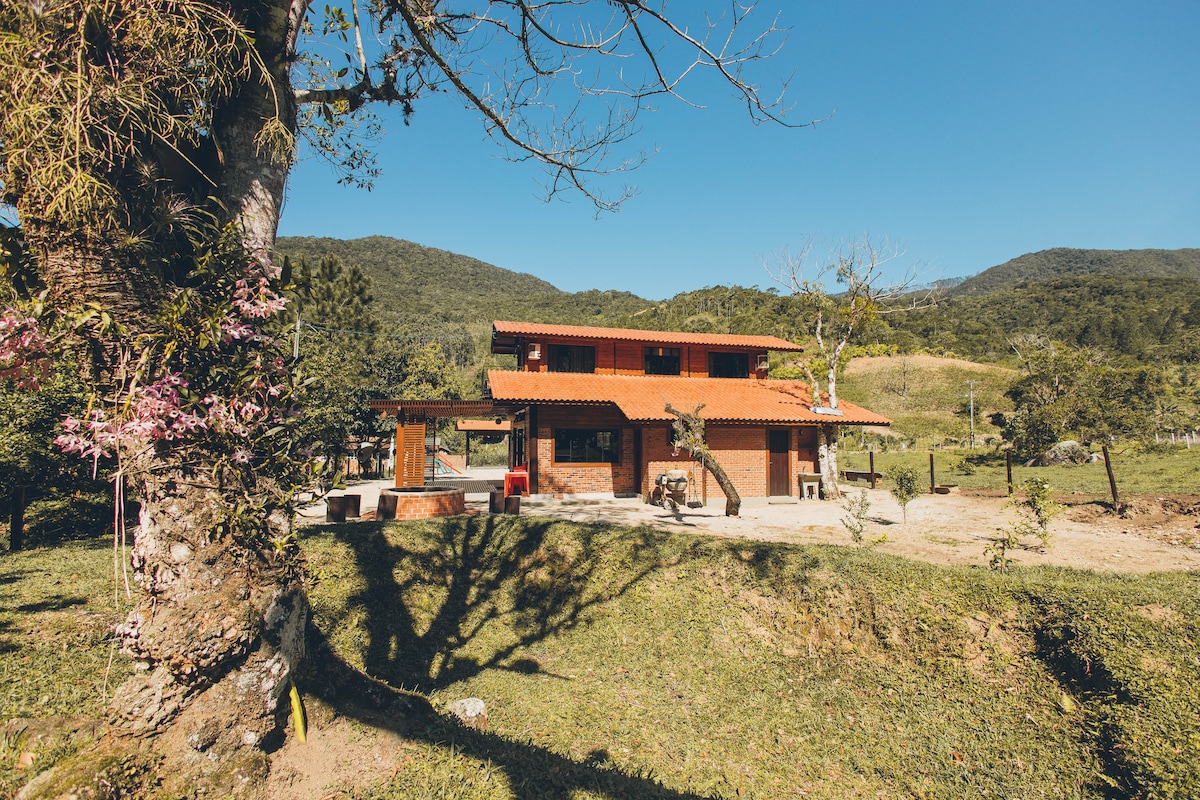 A rustic two-story house with a red-tiled roof is surrounded by lush greenery and mountains. The façade features large windows and a welcoming porch. Nearby, a tree with pink blossoms adds a natural touch, while the open landscape offers a sense of privacy and tranquility.