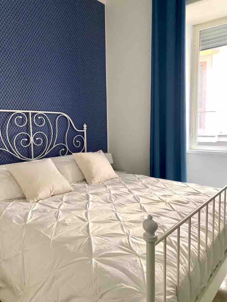 A bright bedroom features a white metal bed frame adorned with a neat, quilted coverlet. Soft, white pillows are placed against the navy blue accent wall. Light enters through a large window, complemented by a set of deep blue curtains.
