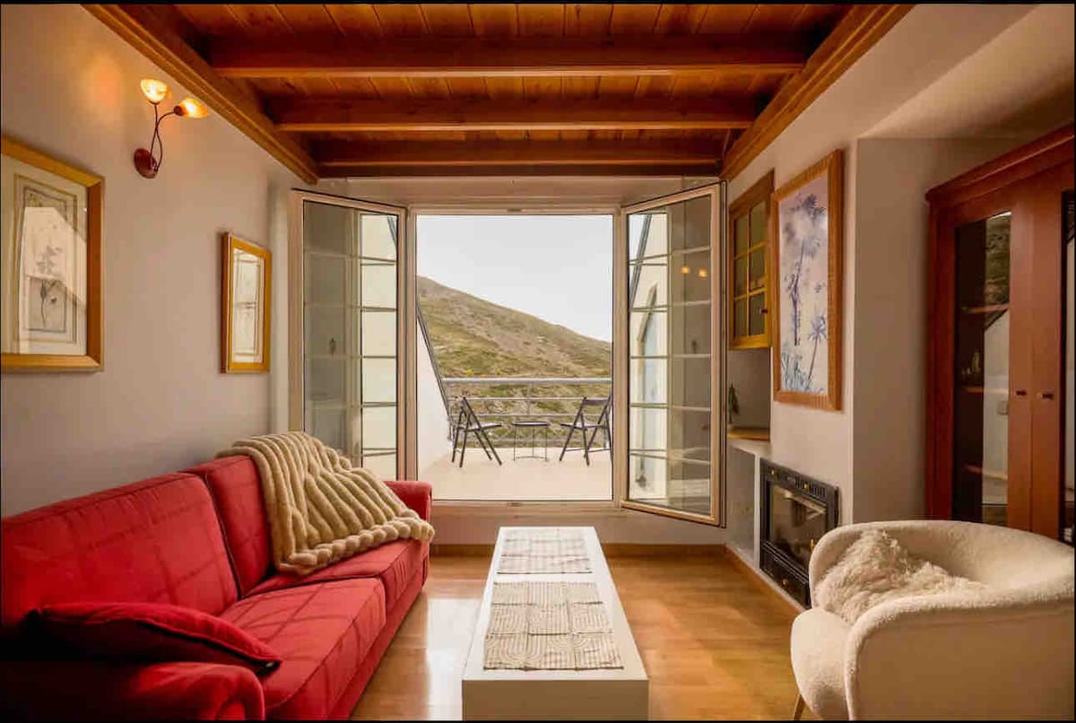 A living area is presented with a red sofa and cozy cream armchair, complemented by a white coffee table. Large glass doors open to a balcony, revealing views of the surrounding mountains. Natural light enhances the warm wooden ceiling and soft furnishings.