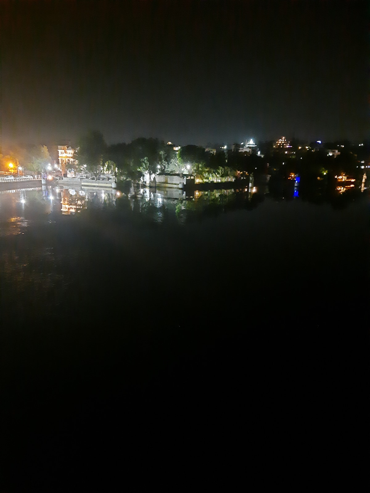 A serene night view of a lakeside landscape is captured, featuring twinkling lights reflecting off the still water. Lush trees line the shore, while illuminated buildings create a gentle glow in the background, enhancing the tranquil atmosphere of the scene.
