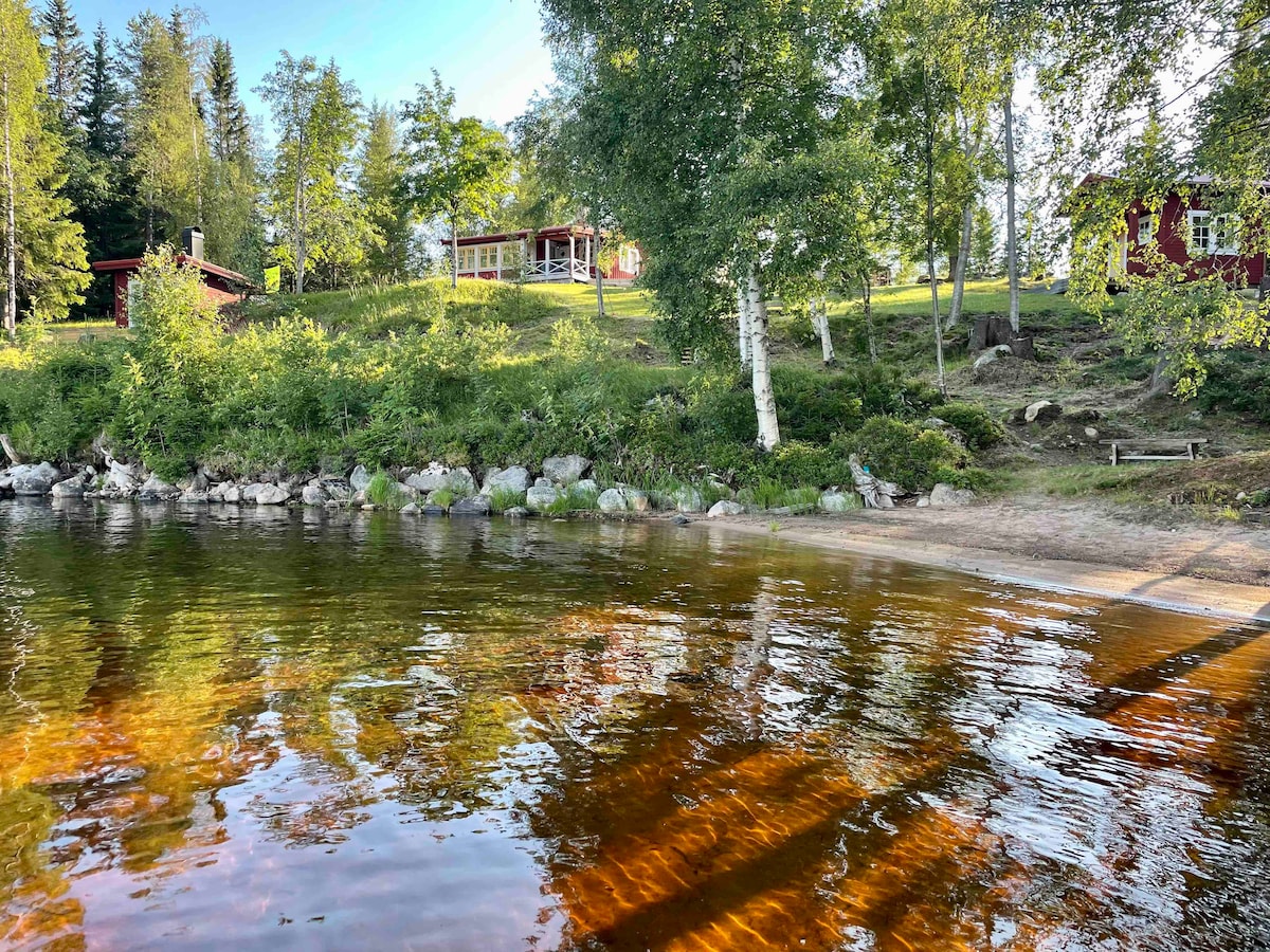 A serene lakeshore is depicted, with clear waters reflecting the surrounding greenery. The sandy beach leads up to a grassy area dotted with trees and the charming red buildings of a cabin complex, providing a peaceful retreat amidst nature.