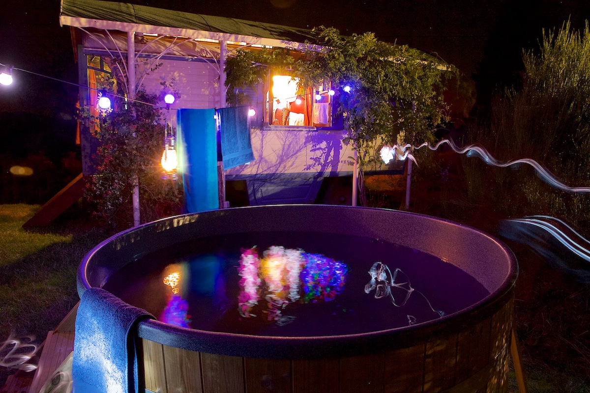 A large Scandinavian hot tub is seen under a starlit sky, with colorful lighting reflecting on the water's surface. The wooden gypsy wagon, adorned with glowing lights, stands nearby, providing a cozy setting for relaxation in nature.