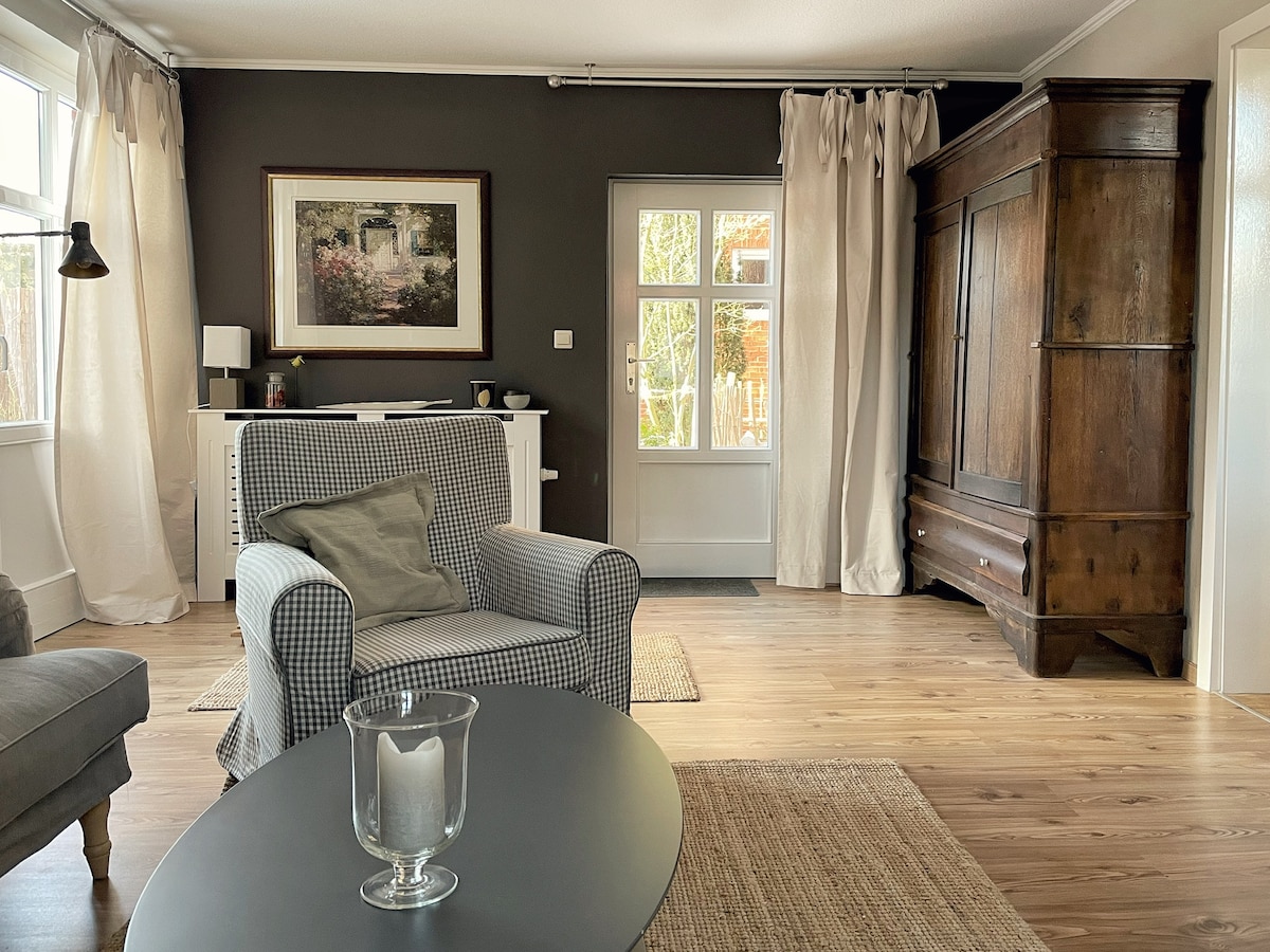 The living room features a cozy seating area with a checked armchair and a gray upholstered chair. A dark wooden cabinet stands against the wall, housing decorative items. Natural light filters through large windows, highlighting the warm wood flooring and subtle textures of the furnishings.