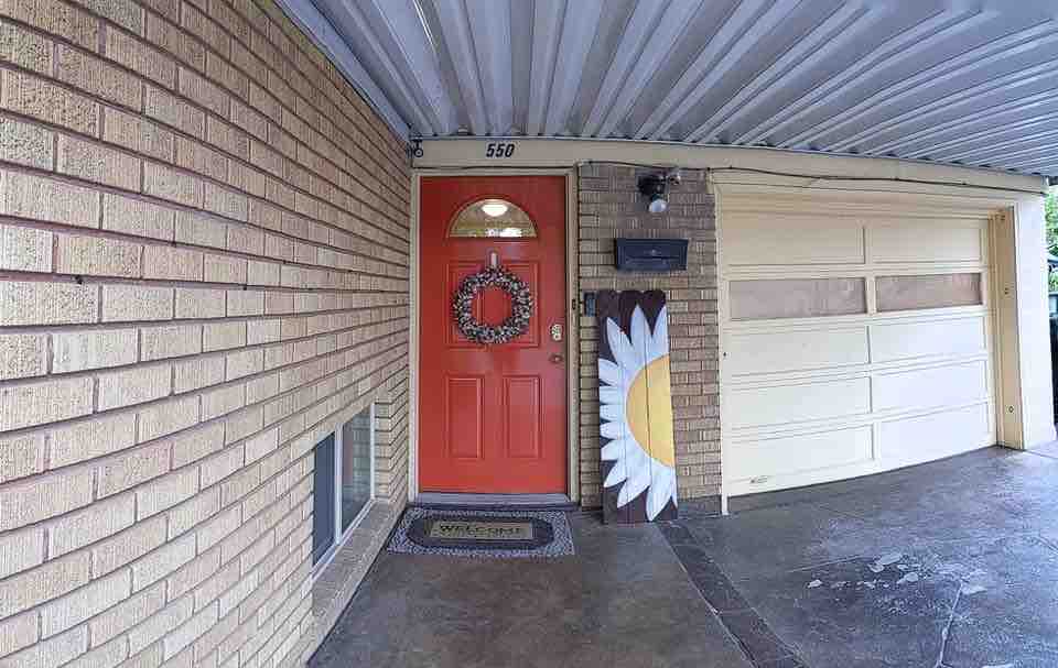 The entrance features a vibrant orange door adorned with a seasonal wreath, welcomed by a decorative doormat. A large, cheerful flower decoration stands beside the door, with a brick exterior and a covered porch providing shelter.
