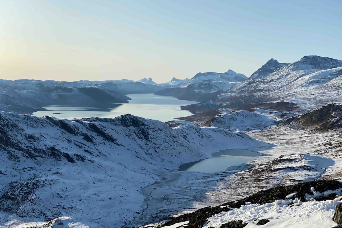 A serene winter landscape is presented, featuring snow-covered mountains and slopes extending towards a calm body of water. The scene captures a tranquil view under clear skies, with distant peaks visible in the background, illustrating the natural beauty of the surrounding area.