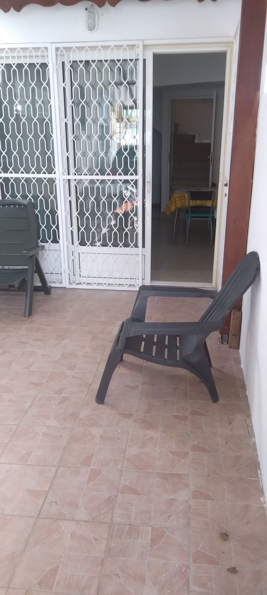 A shaded outdoor area features a single black plastic chair positioned on a tiled floor. In the background, a set of secure white metal doors is seen, leading to an interior space with visible stairs. A dining table adorned with a colorful tablecloth is partially visible.