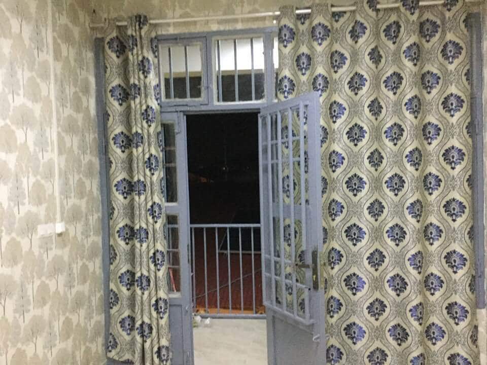 A balcony door with a grill design opens to a view outside, framed by patterned curtains that feature a floral motif. The walls are adorned with a neutral wallpaper, contributing to a cozy ambiance.
