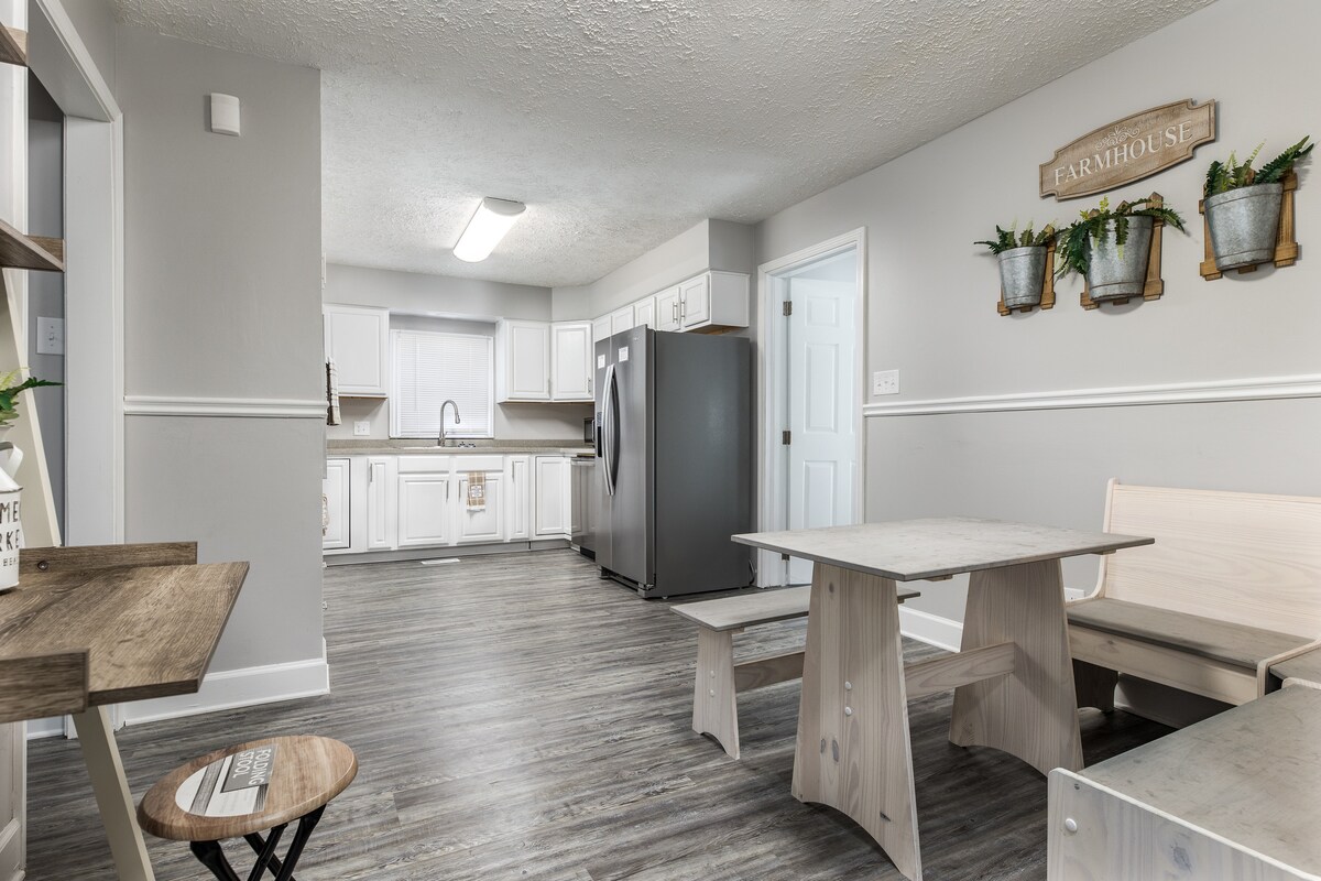A spacious kitchen area features light-colored cabinetry and stainless-steel appliances, including a refrigerator. A wooden dining set, with a bench and round table, is positioned to one side. The flooring is a light wood design, contributing to a clean and open atmosphere.