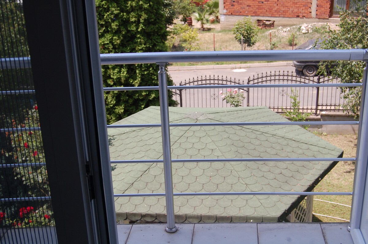 A terrace view captures a green-roofed gazebo situated in a garden, surrounded by greenery and a wrought-iron fence. A pathway is visible beyond the gazebo, leading to additional gardens and parked vehicles.