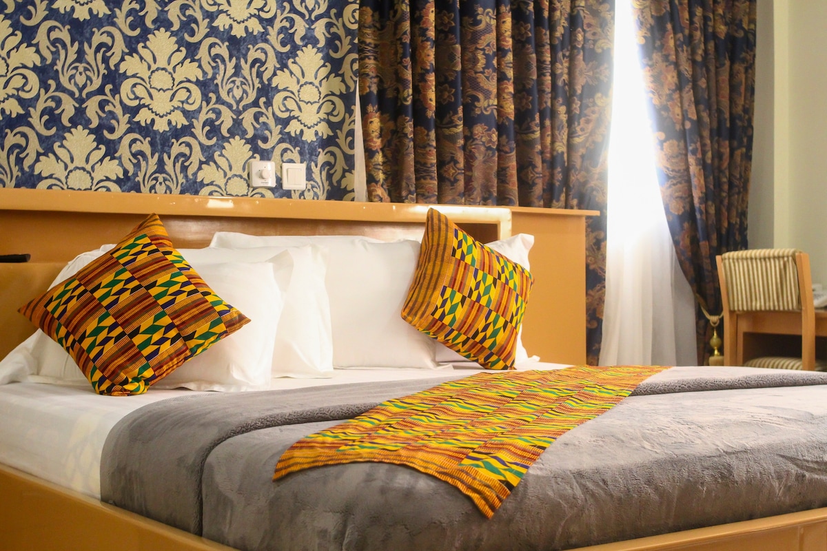 A modern bedroom features a spacious bed adorned with patterned pillows. A colorful throw blanket is placed at the foot of the bed. Elegant curtains frame a bright window, while intricate wallpaper adds a decorative touch to the ambiance.