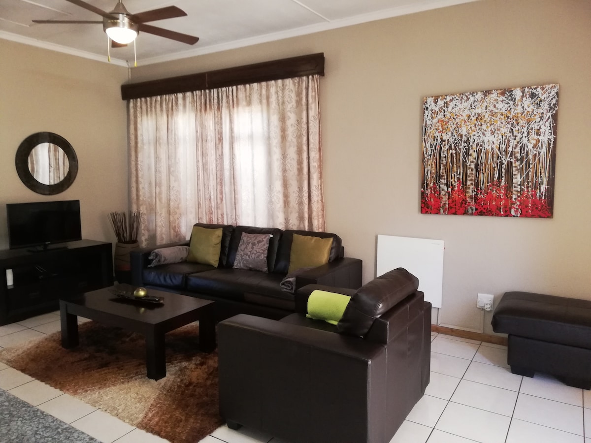 A modern living room features leather couches in dark tones, complemented by colorful cushions. A coffee table rests on a soft area rug. A high-definition TV is positioned against one wall, next to a round mirror. Light filters through sheer curtains, enhancing the relaxing ambiance.