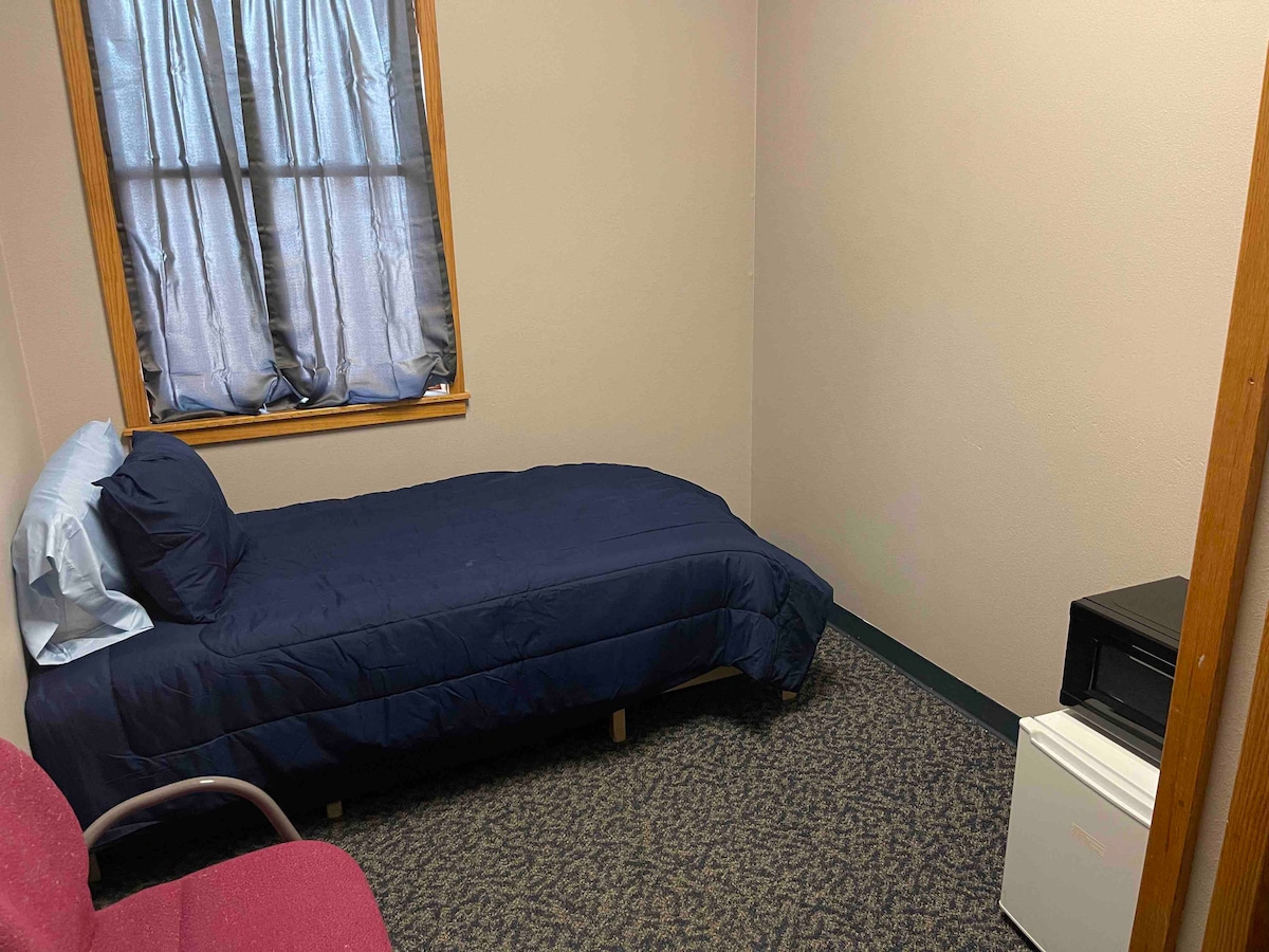 A simple room features a twin bed with a blue comforter and a matching pillow. A small refrigerator and microwave are situated near the foot of the bed. Natural light enters through a window with semi-sheer curtains, illuminating the soft carpet and neutral walls.