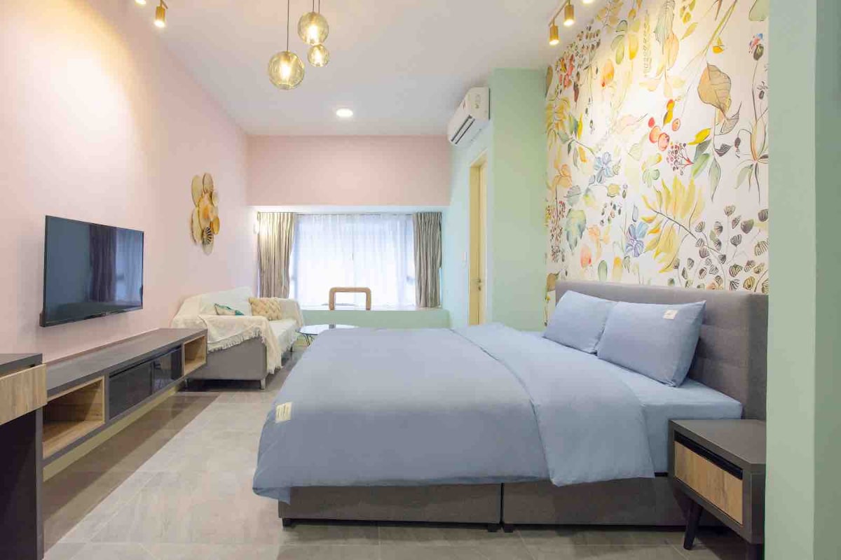 A bright and inviting room features a spacious bed covered with light blue bedding. A floral accent wall adds a touch of nature, while a cozy seating area is visible. Soft lighting fixtures hang from the ceiling, complementing large windows dressed with sheer curtains.