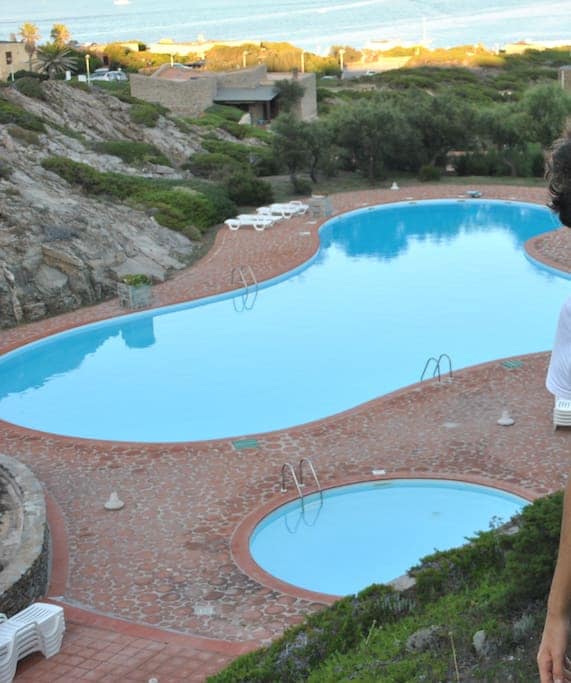 A large swimming pool is surrounded by a stone-tiled terrace with several sun loungers placed around the edge. Lush greenery borders the pool area, while views of the coastline and sea are visible in the background.