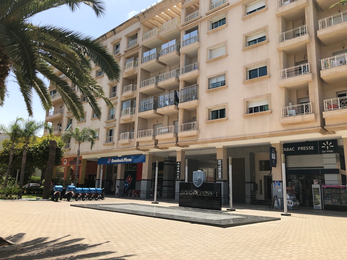 An urban building features a series of balconies with white railings and large windows. Palm trees frame the view, while the ground level displays several storefronts. Delivery scooters are parked nearby, adding to the community atmosphere.