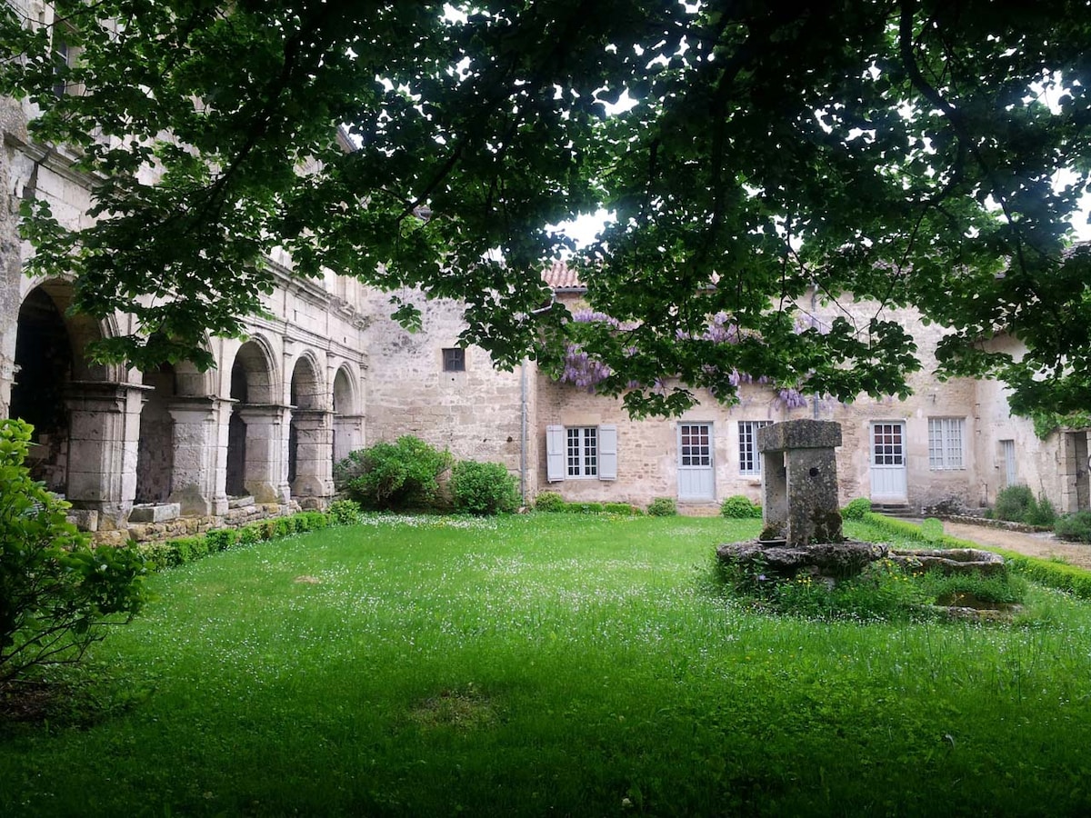 The courtyard showcases a lush green lawn, bordered by stone walls and arches. A plant-covered structure is visible in the background, while a small stone feature stands at the center. Sunlight filters through the tree branches overhead, creating a serene ambiance.