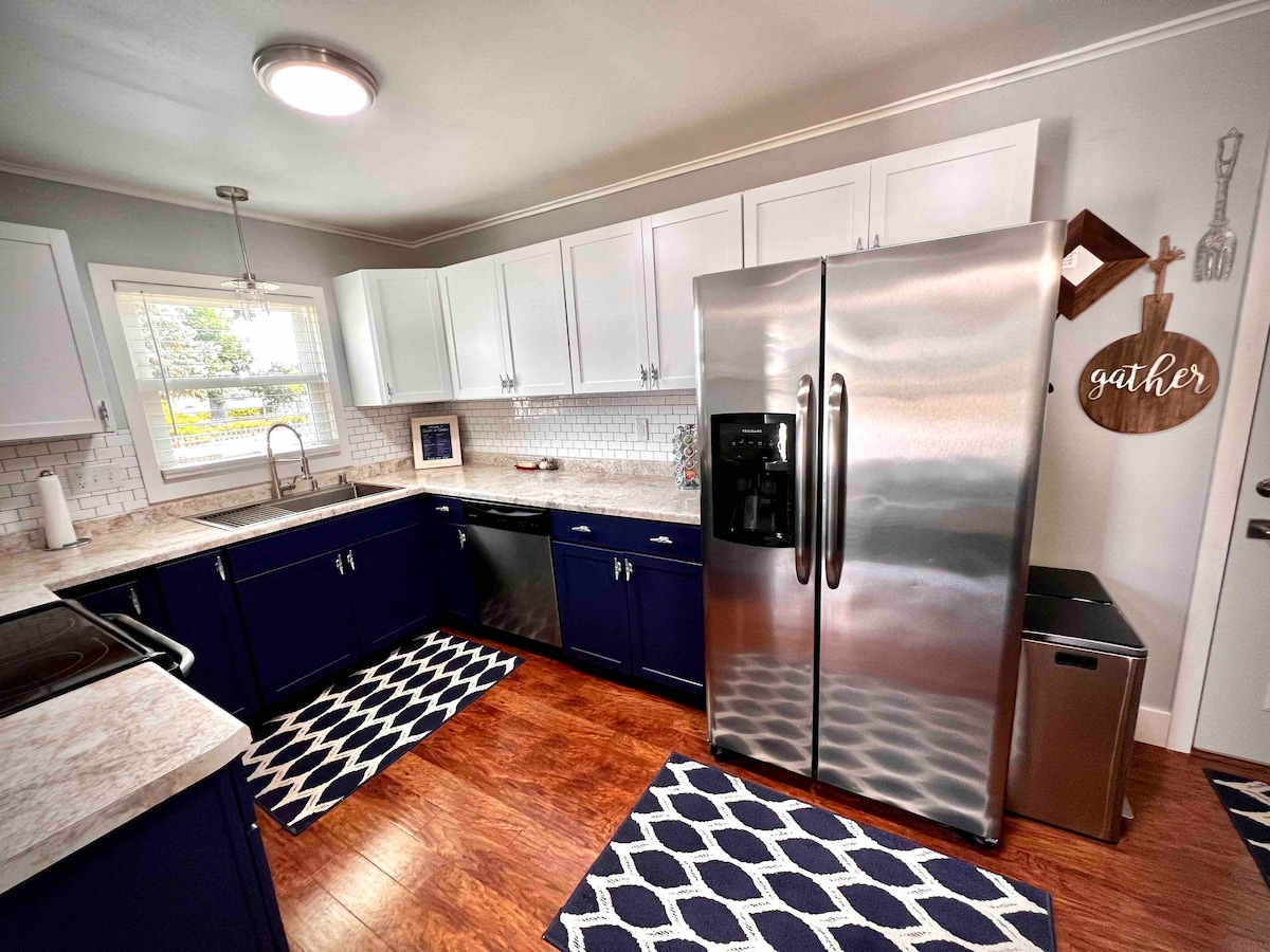A modern kitchen is highlighted by sleek stainless steel appliances, including a large refrigerator. White cabinets contrast with navy blue accents, and a light countertop is complemented by a white subway tile backsplash. A decorative sign reading 'gather' is displayed on the wall.