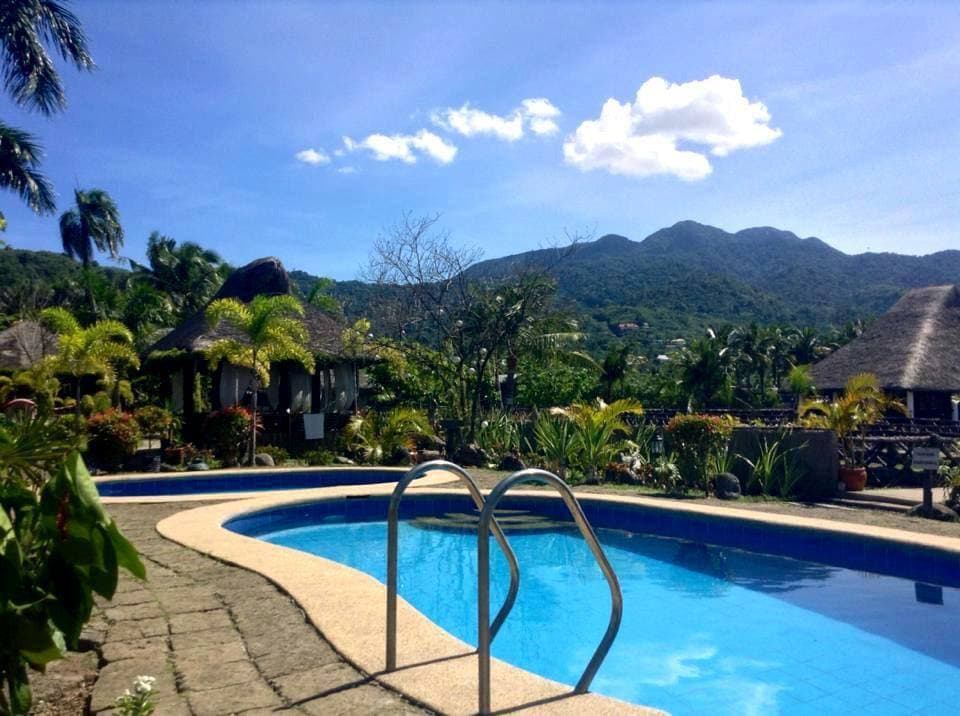 A tranquil swimming pool is framed by lush greenery and vibrant floral arrangements, with a view of the mountains in the background. Thatched-roof cottages are visible nearby, adding a touch of charm to the serene landscape under a clear blue sky.