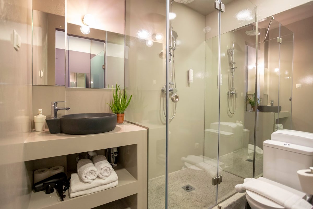 The modern bathroom features a sleek design with a glass shower enclosure. A stylish vessel sink is placed on a compact countertop, accompanied by toiletries. Fluffy towels are neatly arranged, while a small plant adds a touch of greenery.