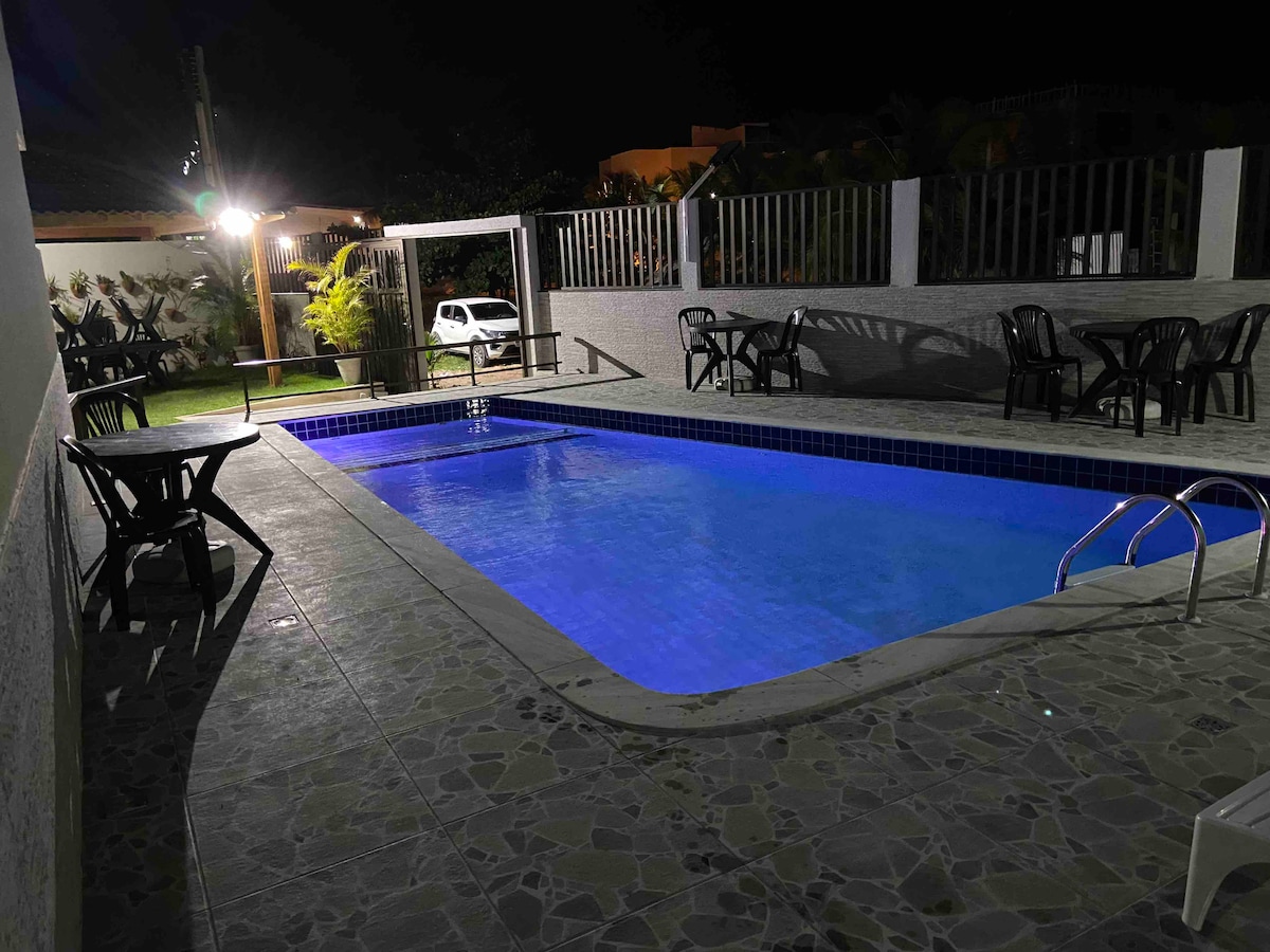 A well-lit pool area features a shimmering blue pool surrounded by a tiled floor. Several black chairs and tables are arranged nearby, creating a space for relaxation. Lush greenery is visible in the background, and outdoor lighting enhances the inviting ambiance.