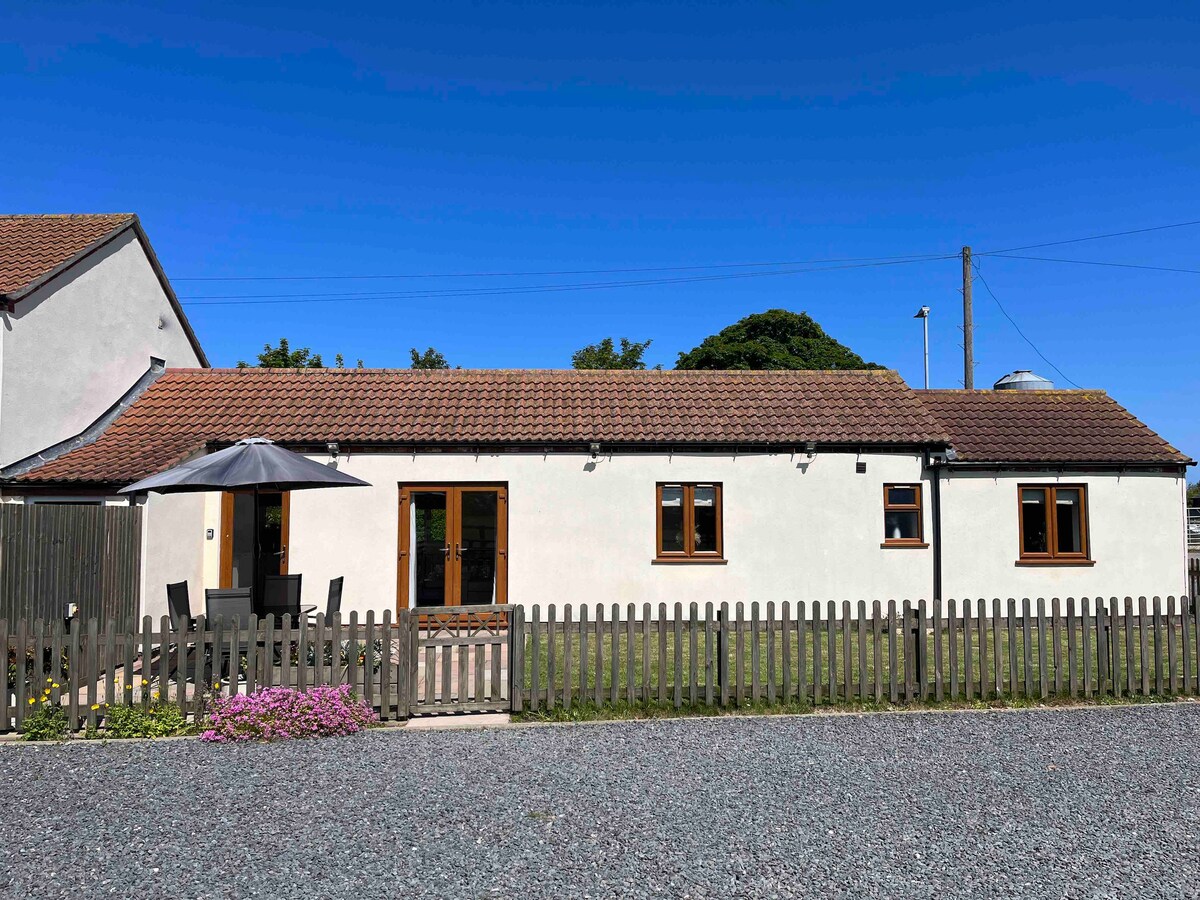 A single-storey barn conversion is showcased with a light-colored exterior and a brown roof. An enclosed garden is visible in front, featuring a seating area under a patio umbrella. A gravel pathway leads to the property, framed by a wooden fence and colorful flower beds.