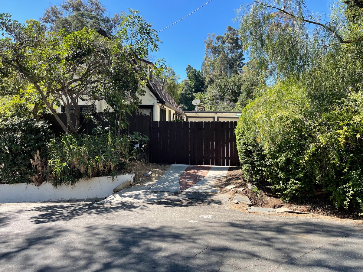A private, gated entrance is showcased with a wooden gate surrounded by lush greenery. The driveway is partially visible, leading up to the cottage, which is nestled among mature trees. Sunlight casts a warm glow over the inviting entrance.