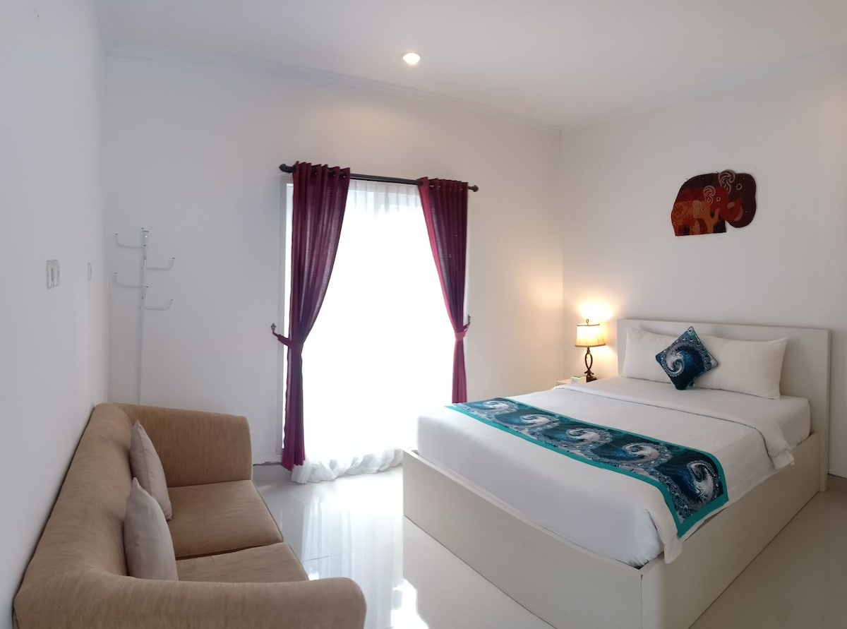 A well-lit room features a spacious bed adorned with decorative pillows. A stylish sofa sits adjacent to a large window, which is dressed in sheer white curtains and dark drapes. A decorative wall piece depicting an elephant adds a cultural touch.