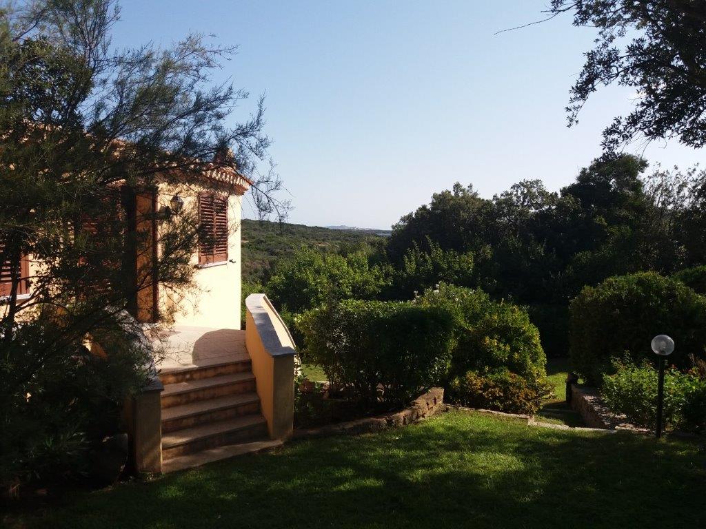 A private villa is surrounded by greenery, featuring neatly trimmed bushes and trees. A set of stone steps leads to the entrance, while distant views of the landscape are visible in the background under a clear blue sky.