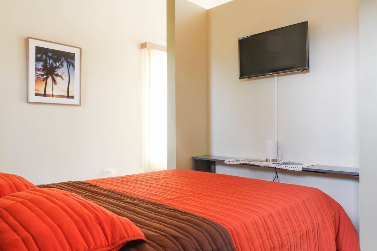 A well-lit studio apartment features a spacious bed dressed in warm orange and brown bedding. A flat-screen television is mounted on the wall above a minimalist desk area, complementing the calming, neutral wall colors. Natural light is provided by a nearby window, enhancing the overall comfort.